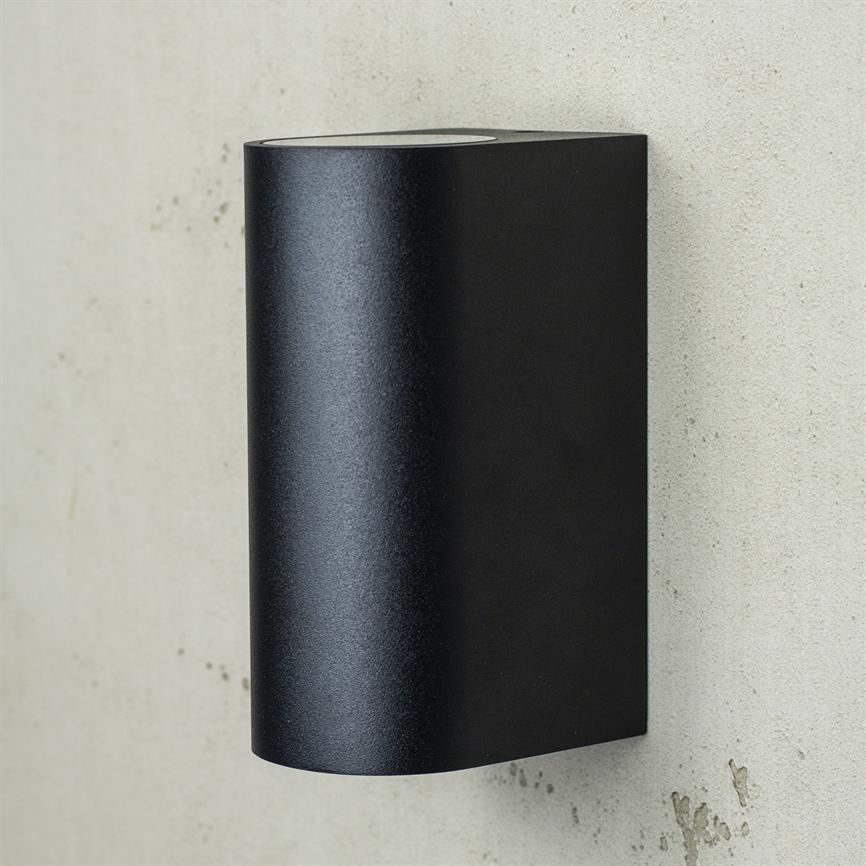 Sort, buet rektangulær væglampe monteret på en lys betonvæg; mat metalfinish med åbning øverst til lys; minimalistisk udendørs design.
