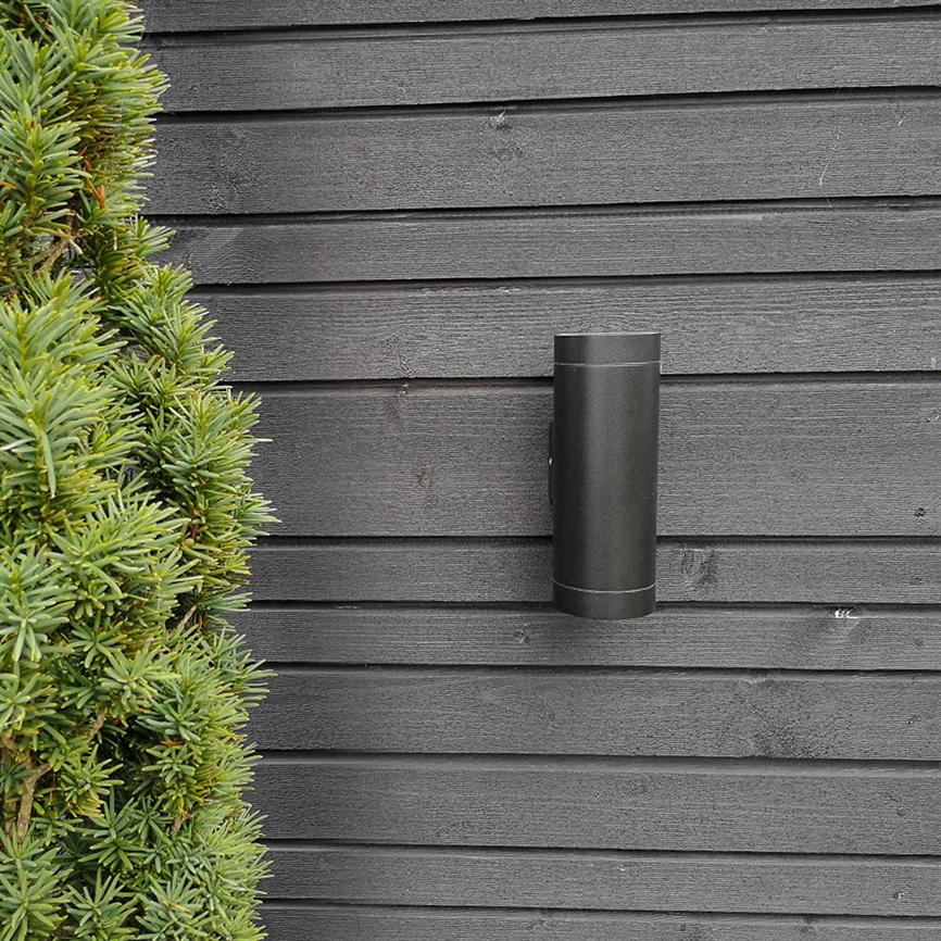 Cylindrisk sort væglampe monteret på mørk, vandret træbeklædning; tæt grønt buskads fylder venstre side, udendørs facade med et minimalistisk, moderne udtryk.
