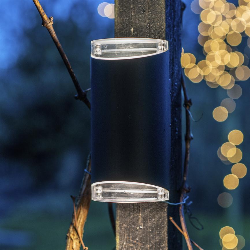 Udendørslampe monteret på en træstolpe, kaster lys opad og nedad; omgivelsen er aftenblå med slyngplanter omkring stolpen og varm, sløret gul lyskæde i baggrunden.
