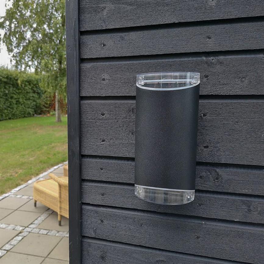 Væglampe, cylindrisk, monteret på sort træbeklædning, lyser ikke; klar plast i top og bund. I baggrunden græsplæne, hæk og flettet havestol ved flisebelagt terrasse.
