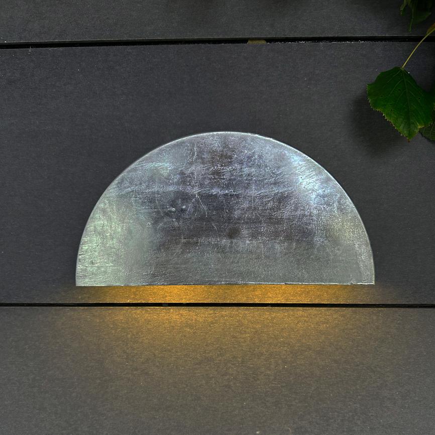 Halvcirkelformet metalplade reflekterer lys og udstråler varmt gult skær nedenunder, monteret på mørk væg med smal sprække og et grønt blad i øverste højre hjørne.