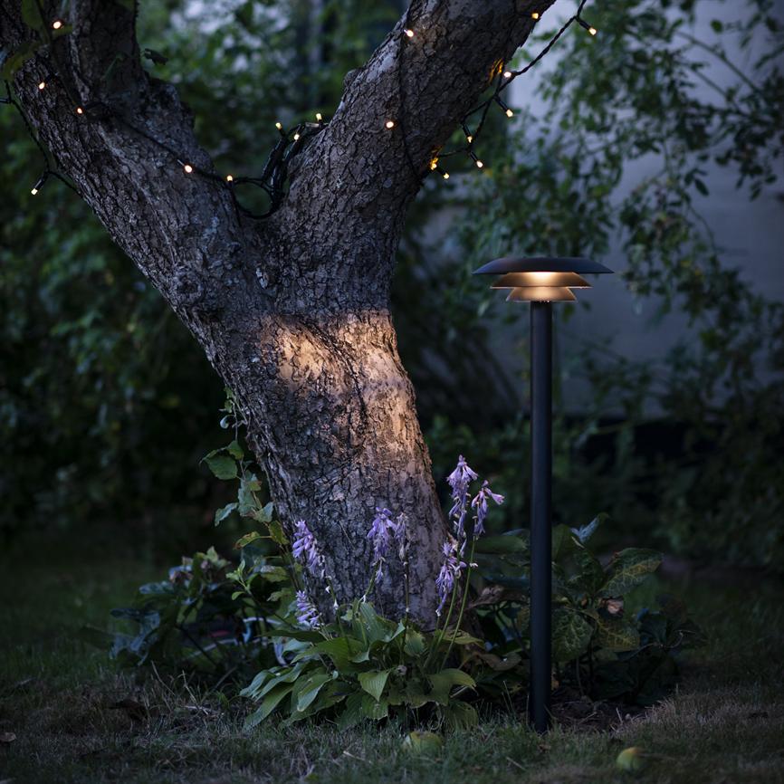 Træstamme oplyses af en lav sort havelampe og små festlige lyskæder, omgivet af lilla hosta-blomster og tæt, mørk haveløv i aftenskumringen.