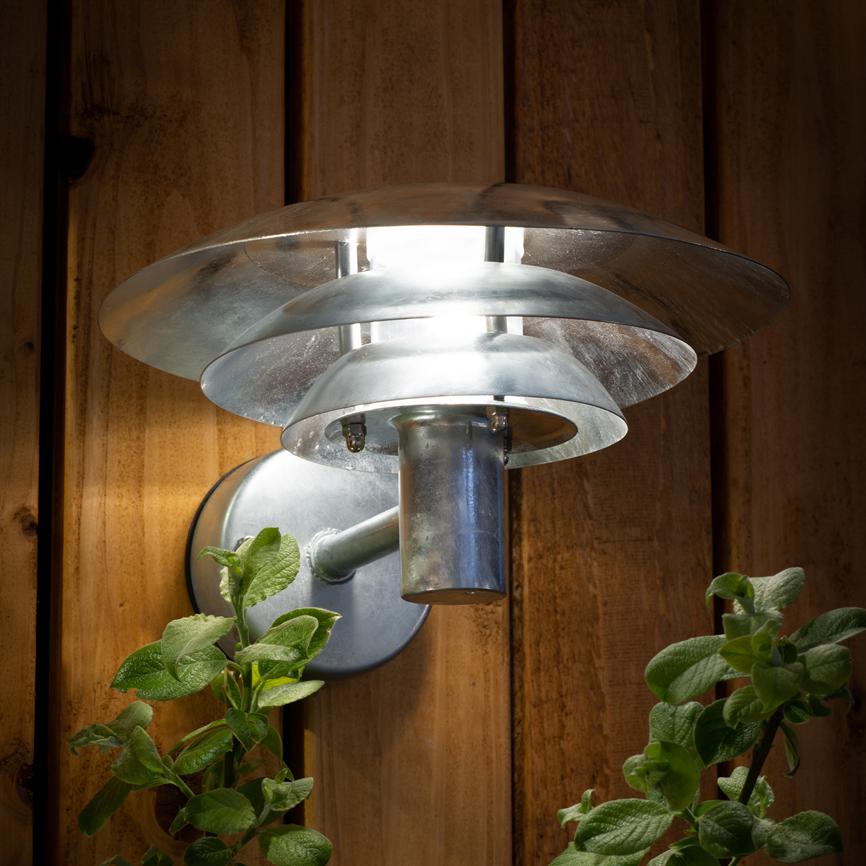 Sølvfarvet metalvæglampe lyser klart nedad og ud, monteret på lodrette træpaneler; grønne planter under lampen fanges af lyset.