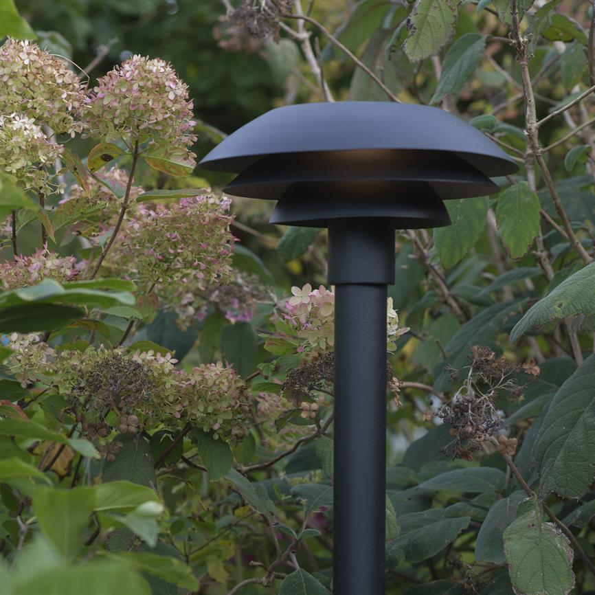 Svampeformet sort havelampe står blandt hortensia og buske, udsender svagt nedadrettet lys gennem lagdelte skærme i en frodig, grøn have.