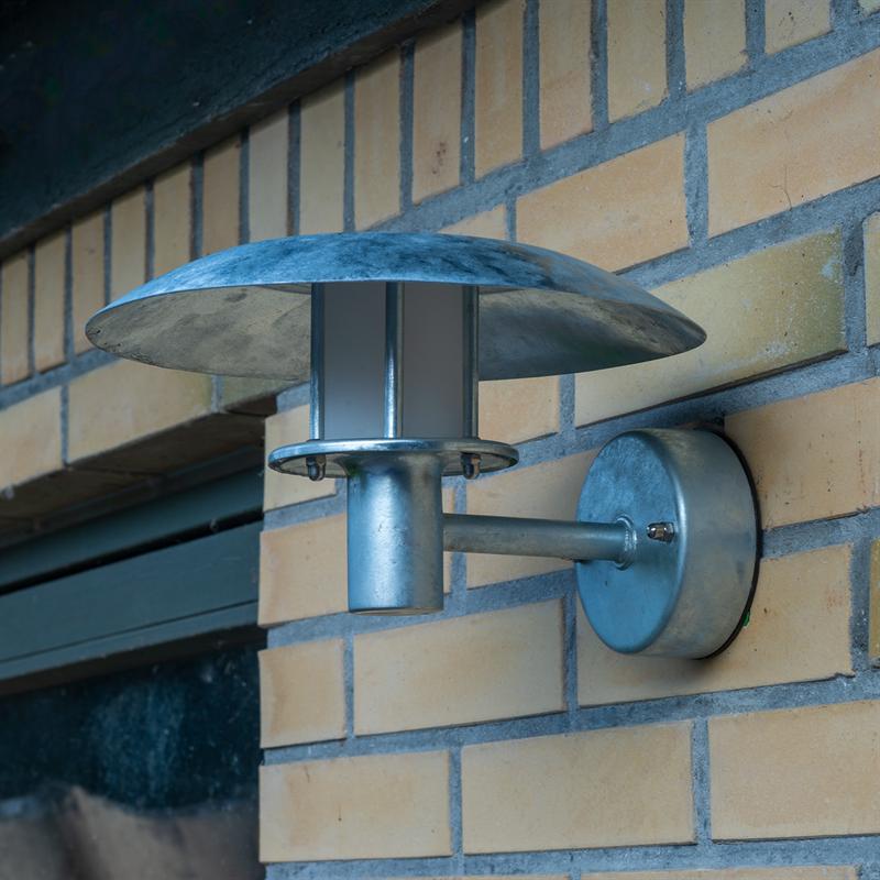Væglampe i galvaniseret metal monteret på en gul murstensfacade; rund skærm hælder over et cylindrisk arm- og lampehus, placeret tæt ved en mørk vindueskarm.