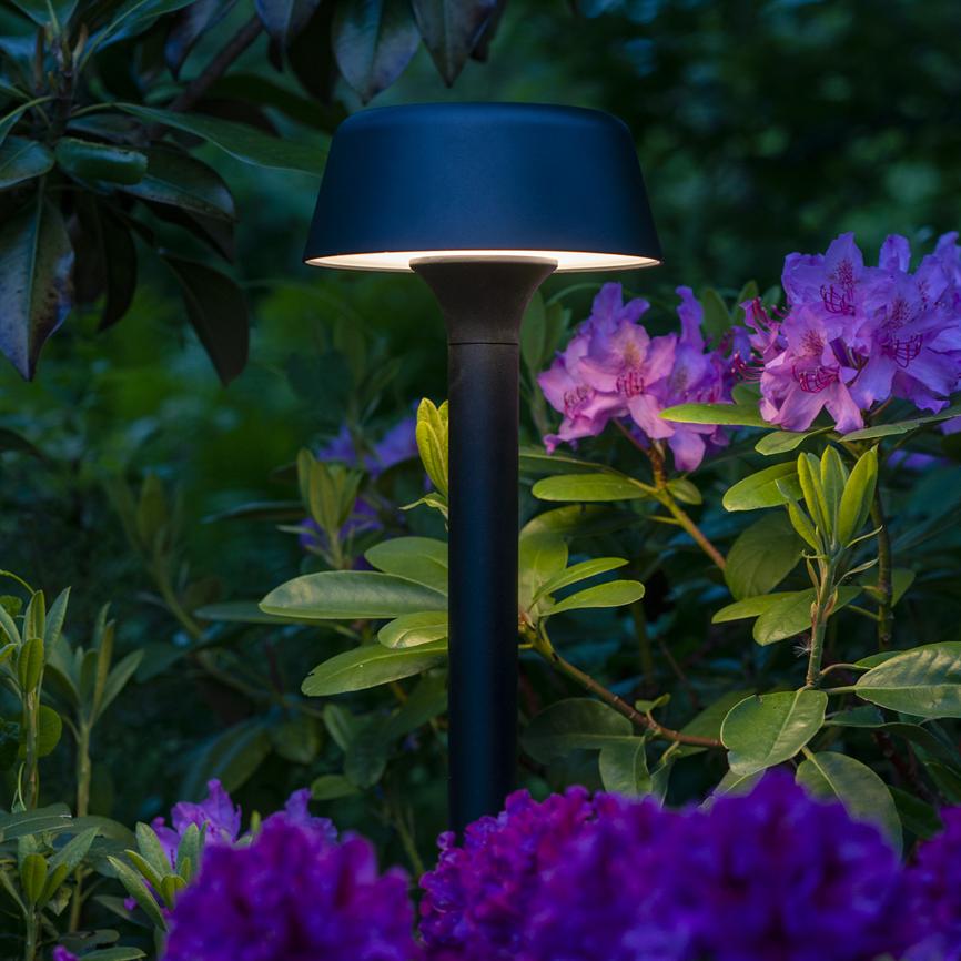 Sort, svampelignende havepælslampe lyser blødt nedad; omgivet af grønne blade og lyse-lilla rhododendronblomster i en frodig, dæmpet aftenhave.
