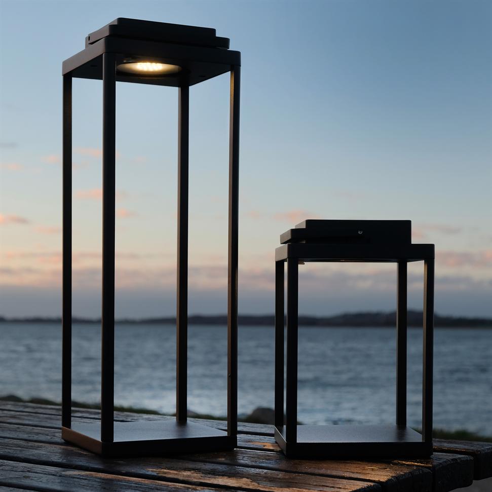 To sorte, åbne metal-lanterner står på et trædæk ved havet; den højere lyser svagt, den lavere er slukket, med kyst og rolig skumringshimmel i baggrunden.