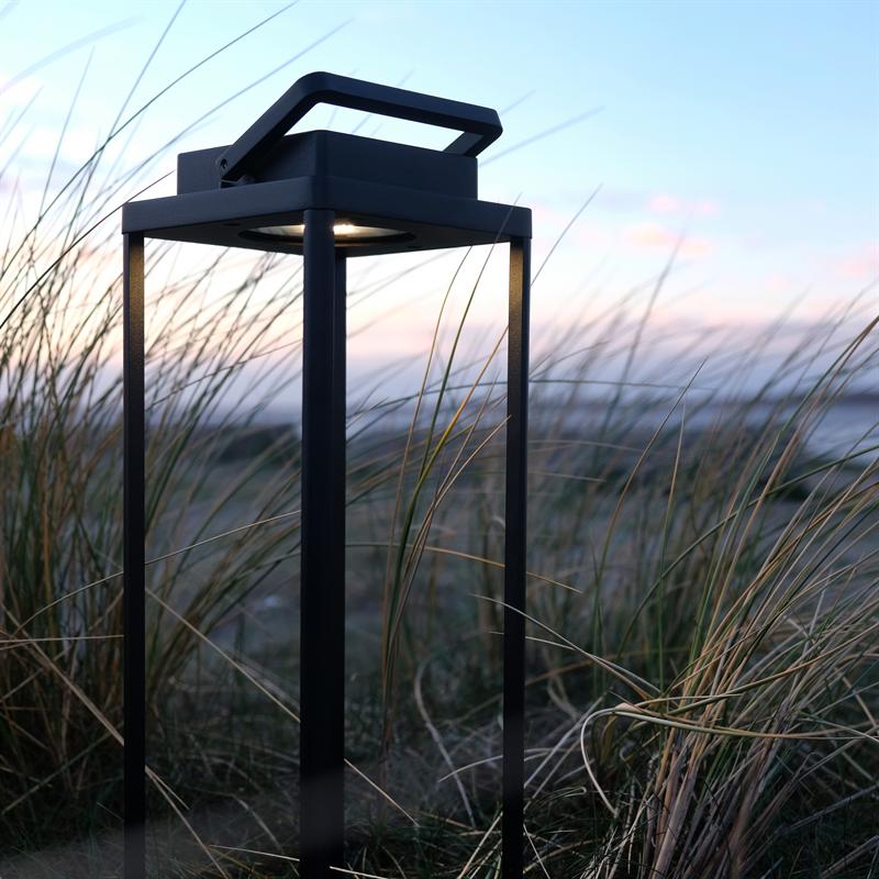 En sort metallanterne står i sandet og kaster svagt lys nedad mellem høje strandgræsser ved skumring, med en blid pastelfarvet himmel og hav i baggrunden.