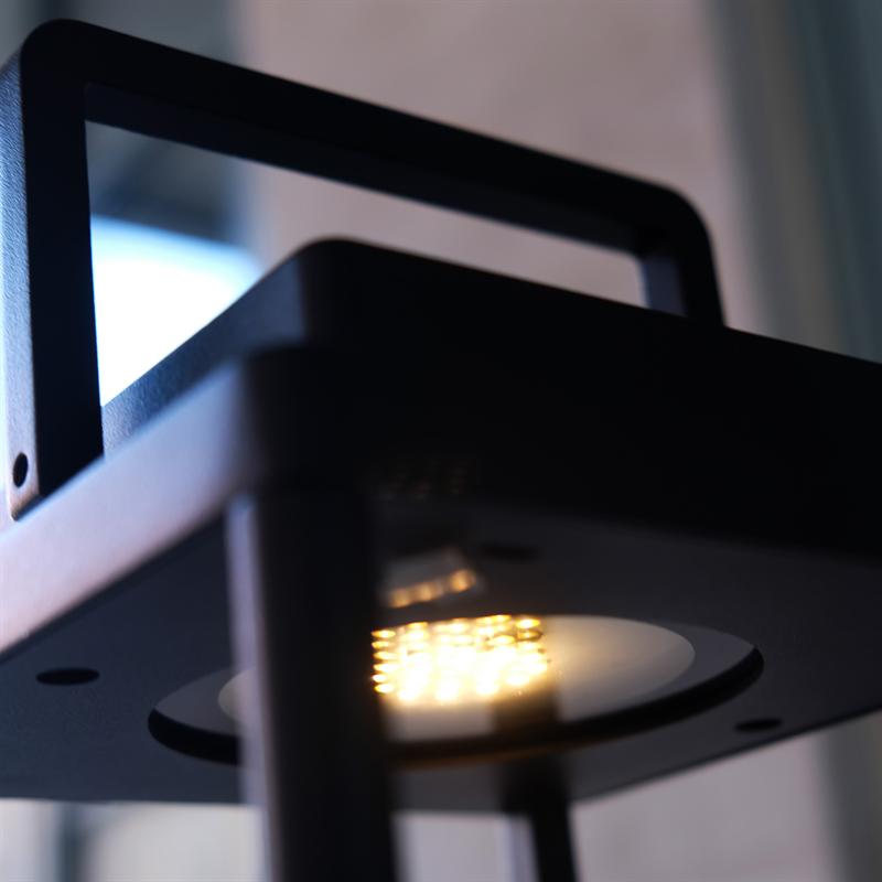 Sort lampekrop med synlige LED-dioder udsender varmt gult lys, monteret i metalramme foran sløret vindue.