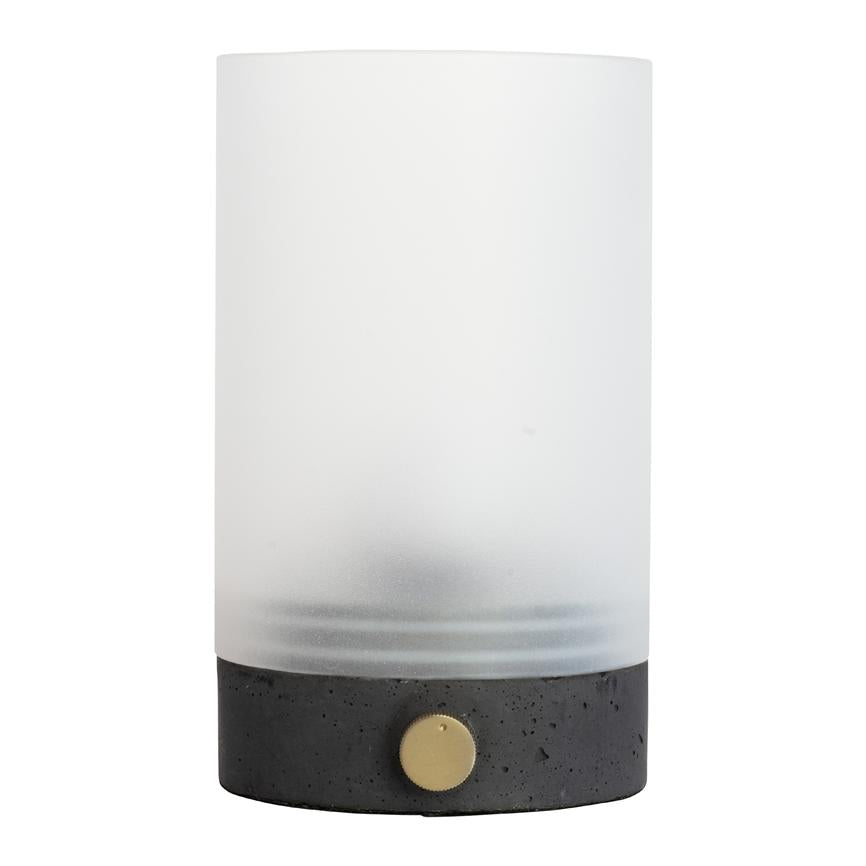 Bordlampe står slukket; frostet glascylinder sidder på en sort betonfod med en lille rund messingknap, placeret mod en neutral hvid baggrund.