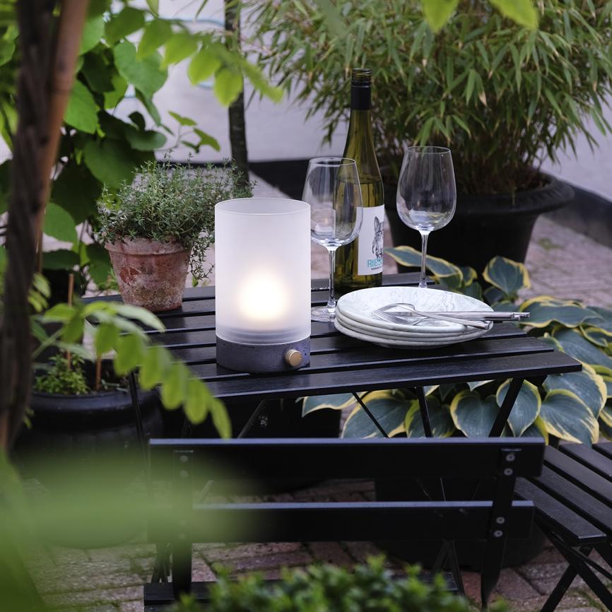 En hvid bordlampe lyser på et lille sort cafébord, omgivet af to vinglas, en vinflaske, en stabel tallerkener og potteplanter på en udendørs terrasse.