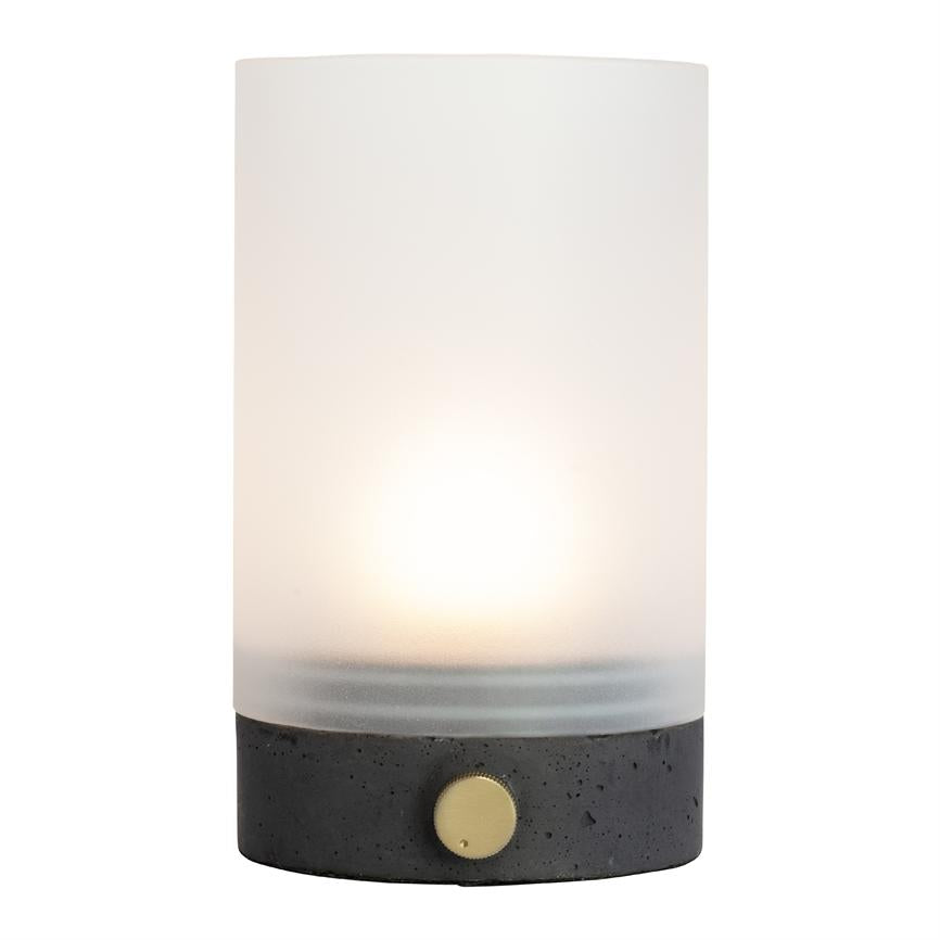 Cylindrisk lampe udsender blødt, varmt lys; mat, frostet glas-skærm over mørk betonfod med guldknap; står på neutral, hvid baggrund.