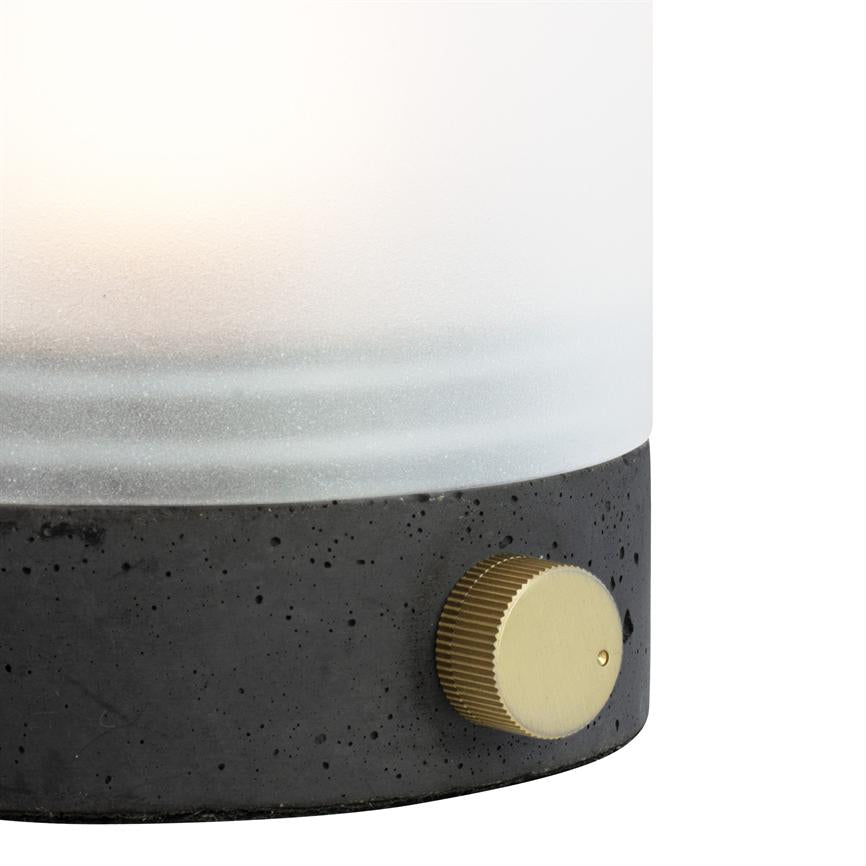 En bordlampe med frostet glaskuppel, der udsender blødt lys; mørk, porøs betonfod hviler på overfladen, og en guldfarvet, rilleret drejeknap stikker ud for regulering.