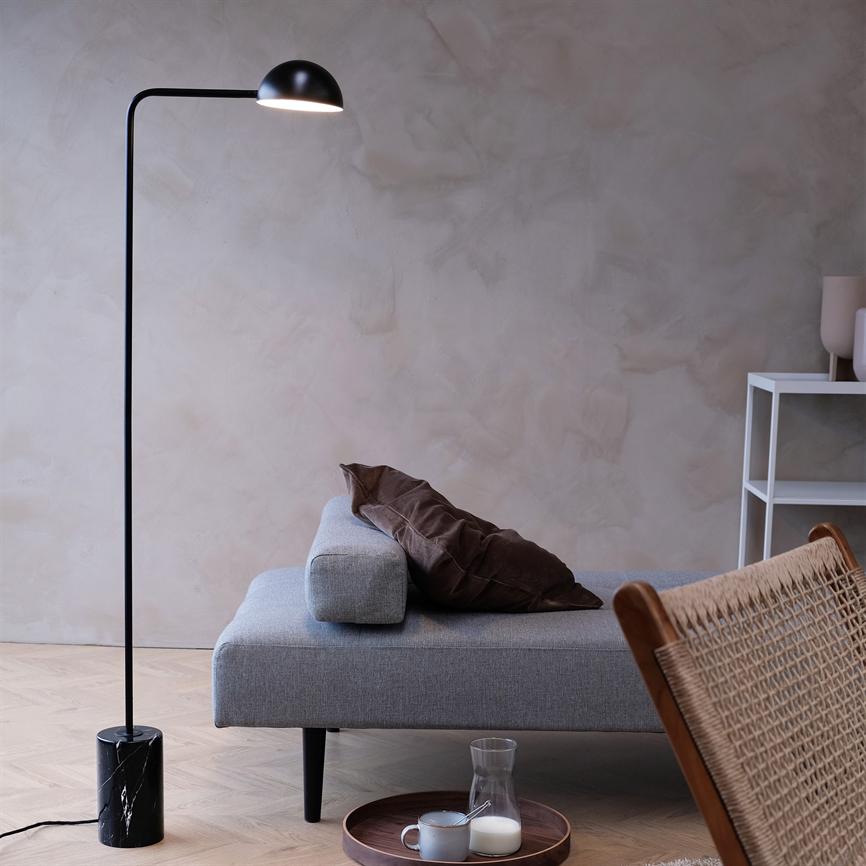 Sort gulvlampe lyser ned over en lav grå sofa; brunt pudebetræk ligger på sofaen, og et rundt træfad med kop og mælkkanne står på lyst trægulv i en minimalistisk stue.