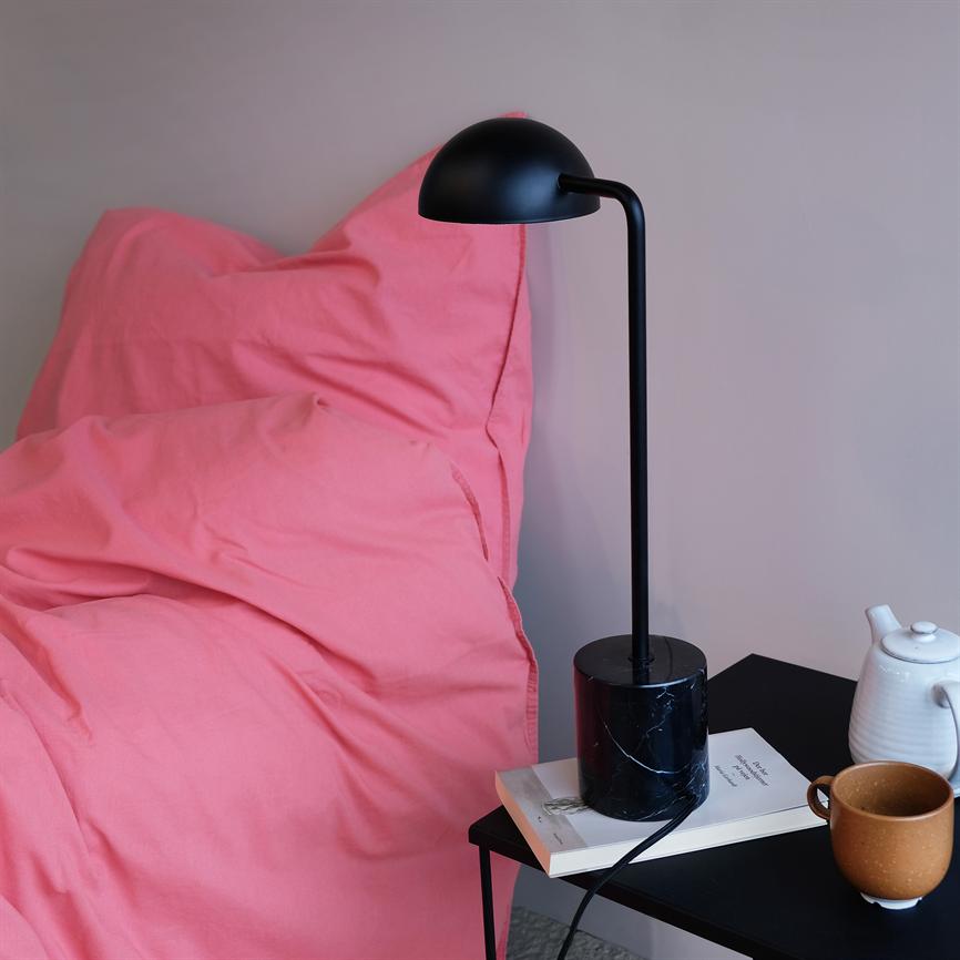 En sort bordlampe står på et sort natbord ved en lyserød dyne; en kop, en tekande og en bog ligger ved siden af i et roligt soveværelse.