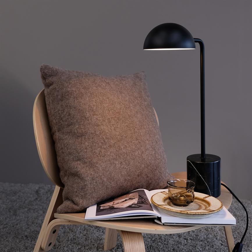 Pude hviler imod en træstol; et åbent magasin, en glas kop på en tallerken ligger på sædet; en sort bordlampe kaster varmt lys i et roligt, gråt rum.