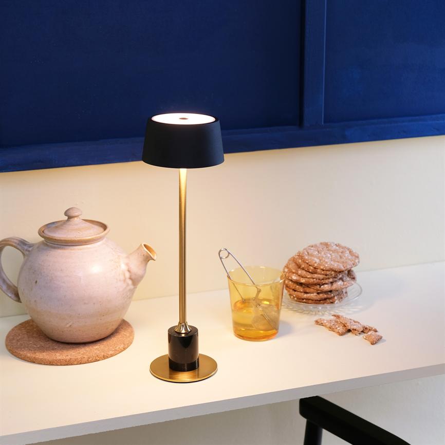 Bordlampe lyser blødt over et hvidt bord; ved siden af står en keramisk tekande på en korkmåtte, et glas med teskeer og en stak småkager foran en mørkeblå væg.