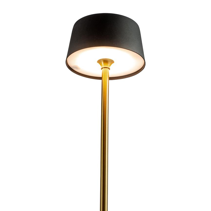 Guldfarvet standerlampe med sort cylindrisk skærm, tændt og kaster blødt, varmt lys nedad; enkel, moderne stil mod en neutral, hvid baggrund.