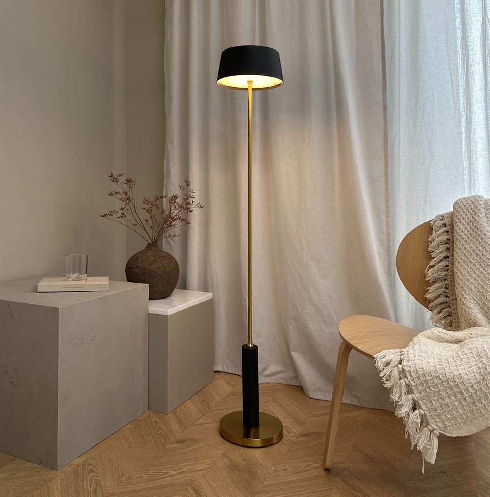 En slank gulvlampe i sort og messing står tændt og kaster varmt lys; den står ved en træstol med en strikket plaid, foran lette gardiner og en keramikvase på betonplinter.