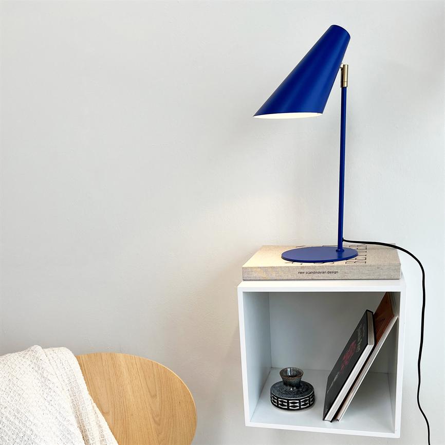 Blå lampe lyser ned på en bog, placeret på en hvid væghængt kassehylde; hylden rummer en lille keramikskål og bøger, ved siden en træstol med hvidt tæppe.
 Tekst på bogen: "new scandinavian design"