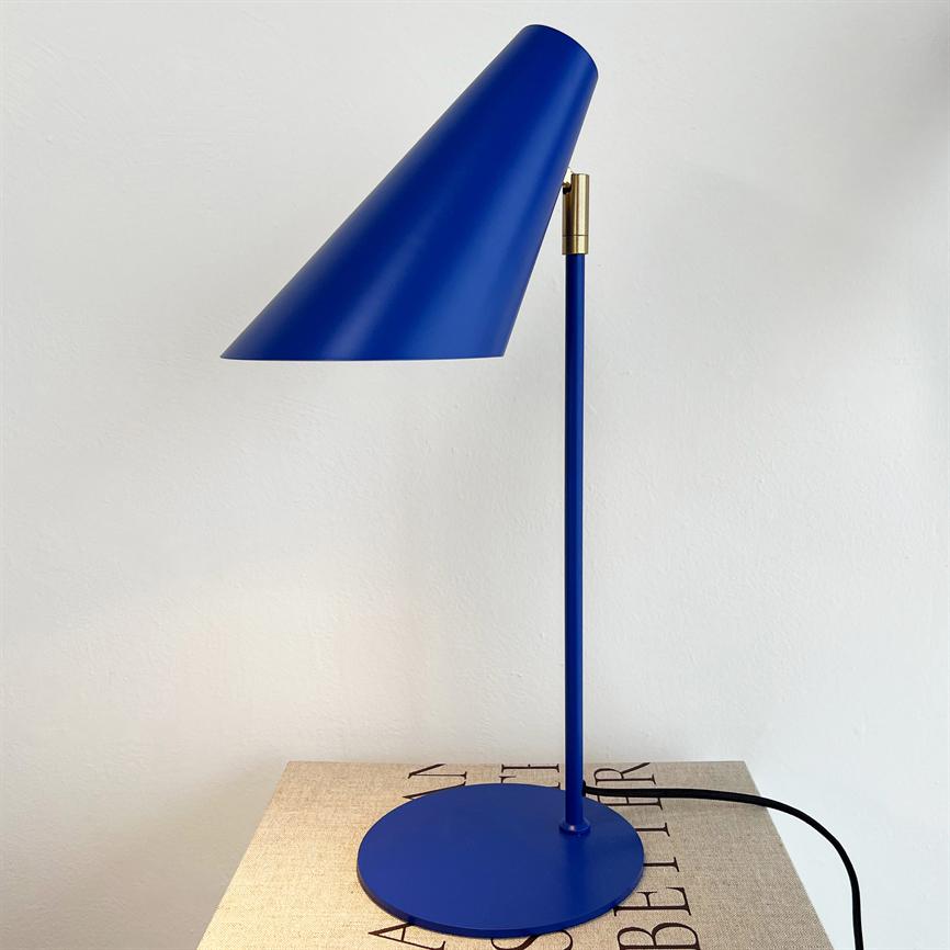 En blå metal skrivebordslampe vinkles med skærmen nedad, placeret på en beige bog/æske foran en hvid væg. Tekst: "AS BETTER" (delvis skjult).