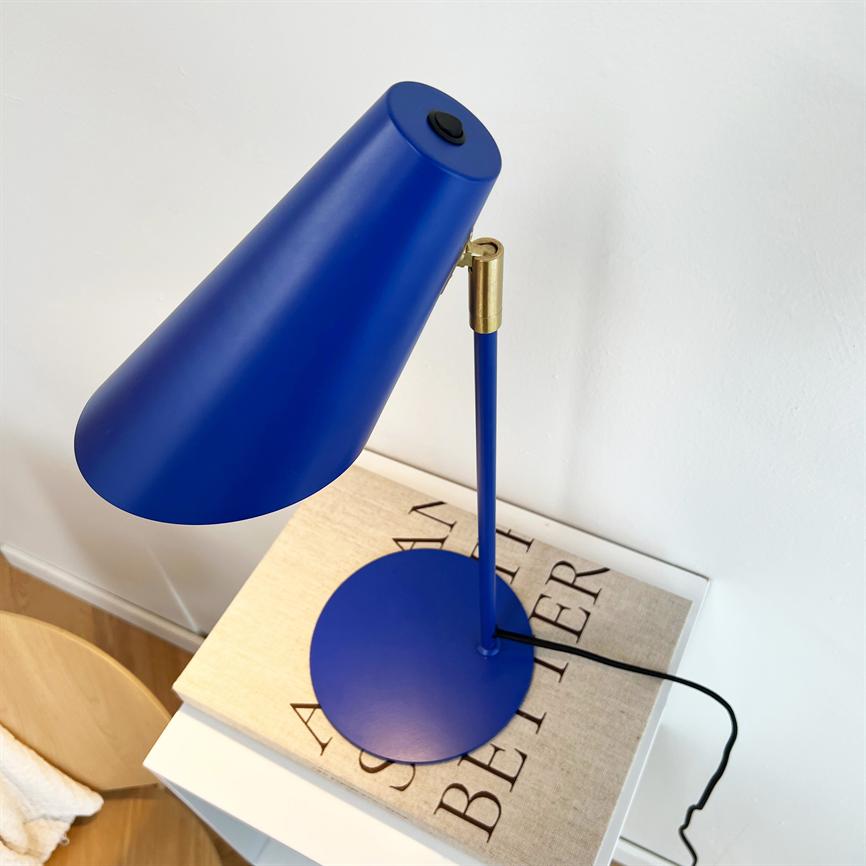 Blå bordlampe hælder fremad over en bog på et hvidt natbord foran en hvid væg; sort ledning løber ud over bordkanten. 
 Tekst på bog: "AS A M BETTER"