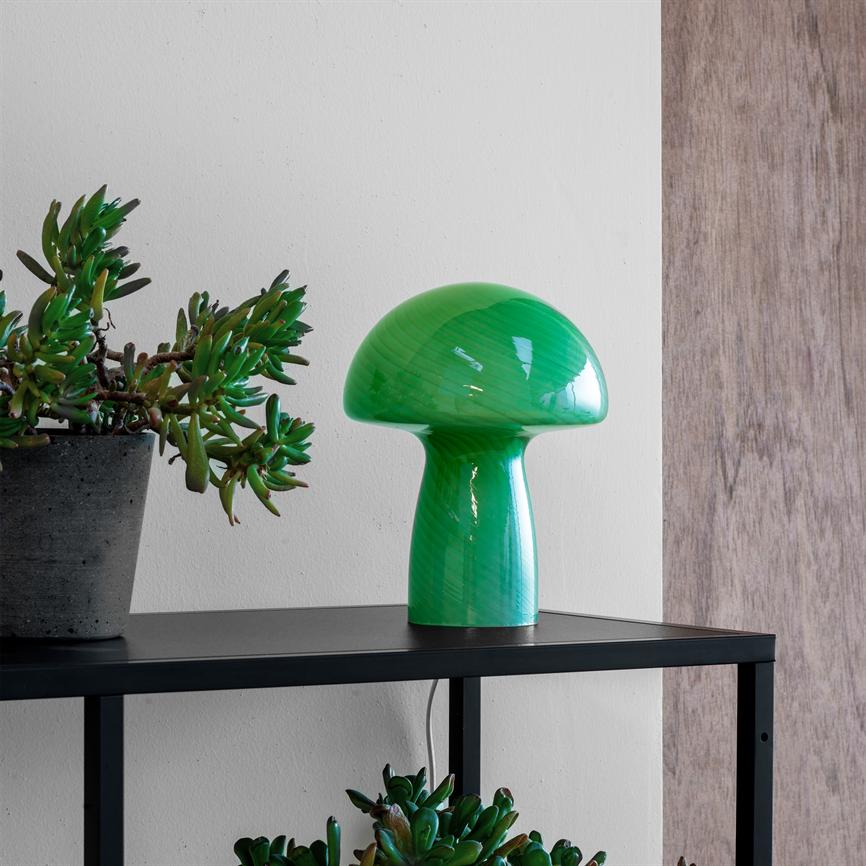 Grøn svampeformet lampe står på en sort metalhylde, flankeret af sukkulenter i betonpotter, foran en hvid væg med en træplade til højre.