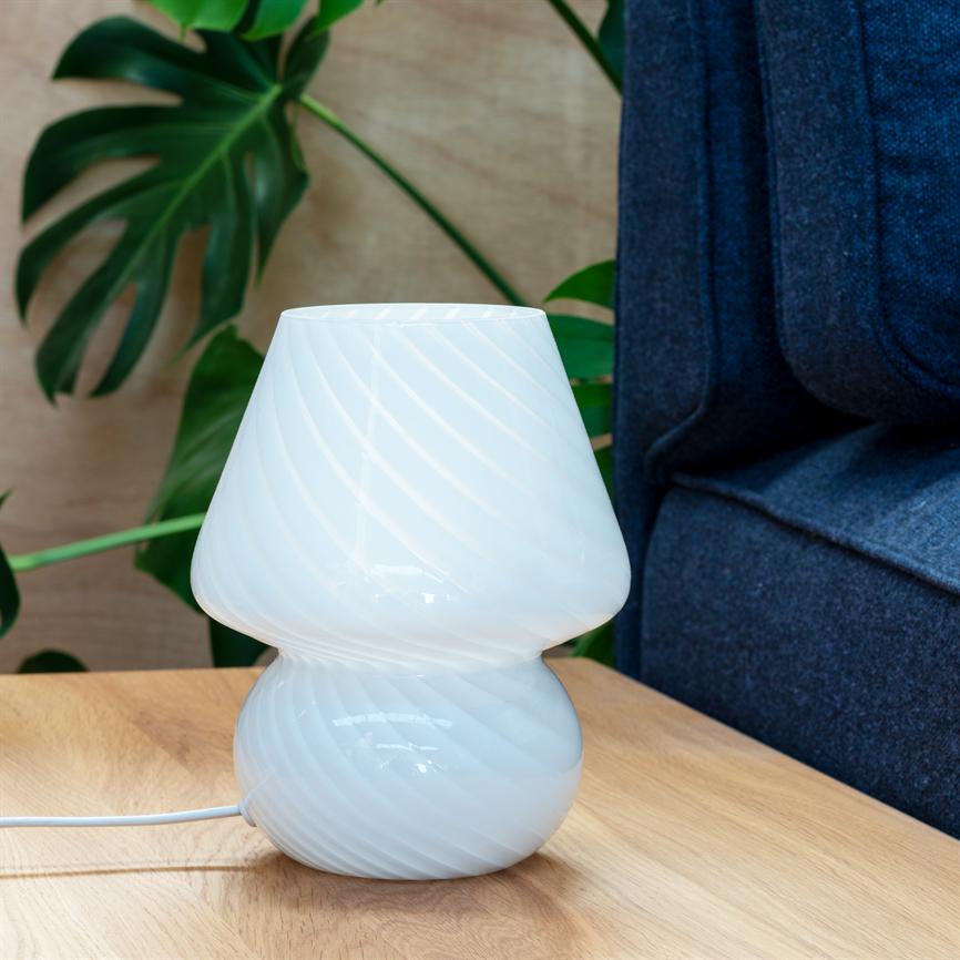 Hvid, spiralmønstret glasbordlampe står på et lyst træbord; den er slukket, placeret ved en mørkeblå sofa og grønne stueplanter i et hyggeligt stueinteriør.