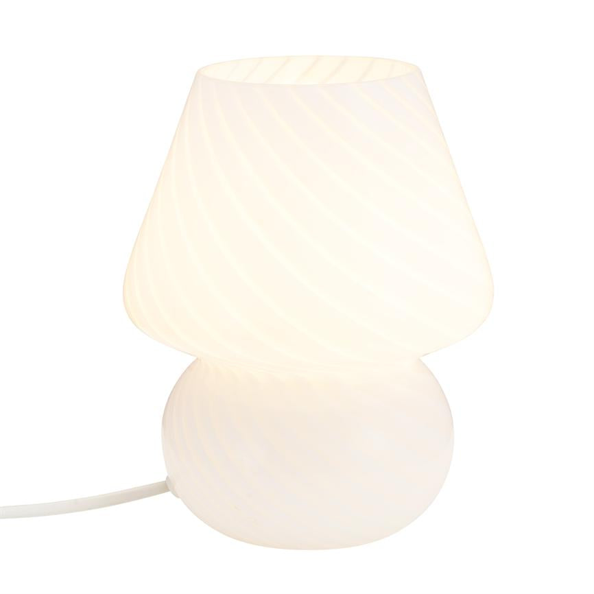 Bordlampe udstråler blødt, varmt lys; rund base og bred, let mønstret skærm set mod ensfarvet hvid baggrund, med hvid strømkabel synligt.