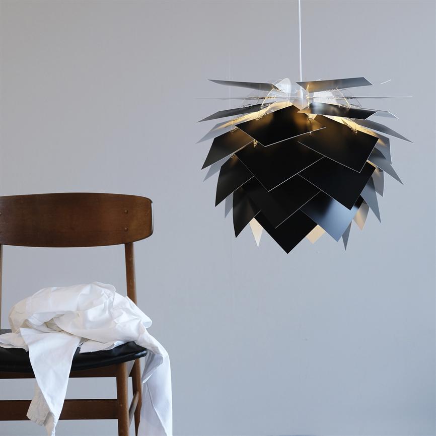 En lagdelt sort pendellampe svæver tændt foran en grå væg; til venstre står en træstol med en hvid skjorte draperet over sædet.
