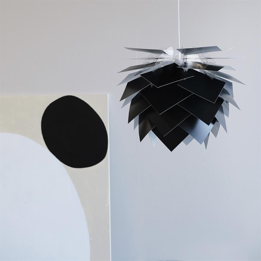 En sort-sølv lagdelt pendellampe hænger fra loftet; dens overlappende metalplader danner en koglelignende form. I et lyst, minimalistisk rum ved siden af et abstrakt maleri med stor sort cirkel.