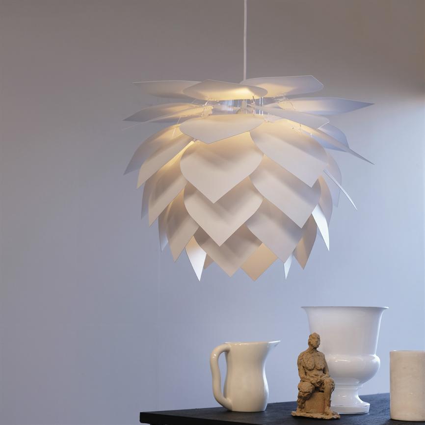 Hængelampe af overlappende blade hænger og udsender varmt, diffust lys over et mørkt bord med en hvid kande, hvid vase og en lille siddende lerfigur foran en lys væg.