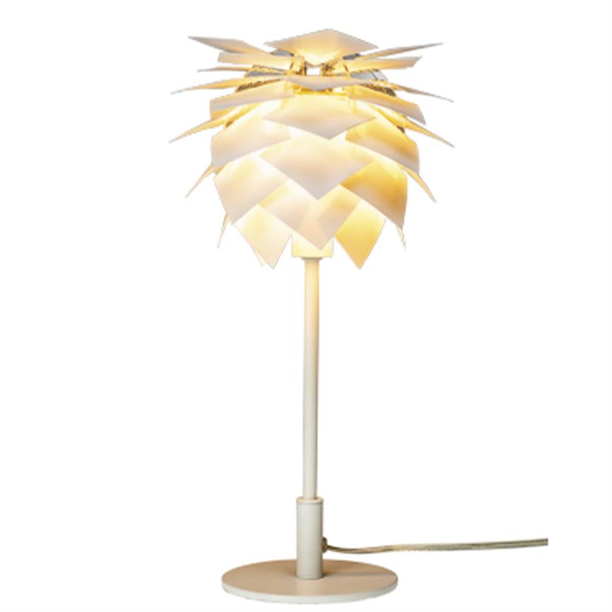 En hvid bordlampe med skærm af overlappende, bladlignende plader udsender varmt, gyldent lys; monteret på en slank stang og rund fod, placeret mod en neutral, lys baggrund.