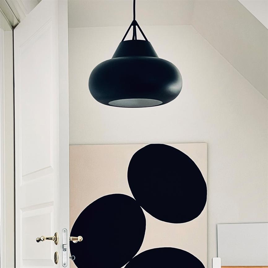 Sort hængende lampe svæver over et lærred med tre store sorte cirkler, placeret mod en hvid væg i et lyst rum; en delvis åben hvid dør med messinghåndtag i forgrunden.