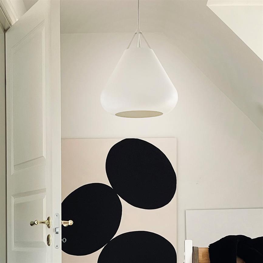 En hvid pendel hænger midt i rummet over et stort, lyst lærred med tre overlappende sorte cirkler; en åben dør og en sengegavl er synlige i et minimalistisk, lyst loftsrum.