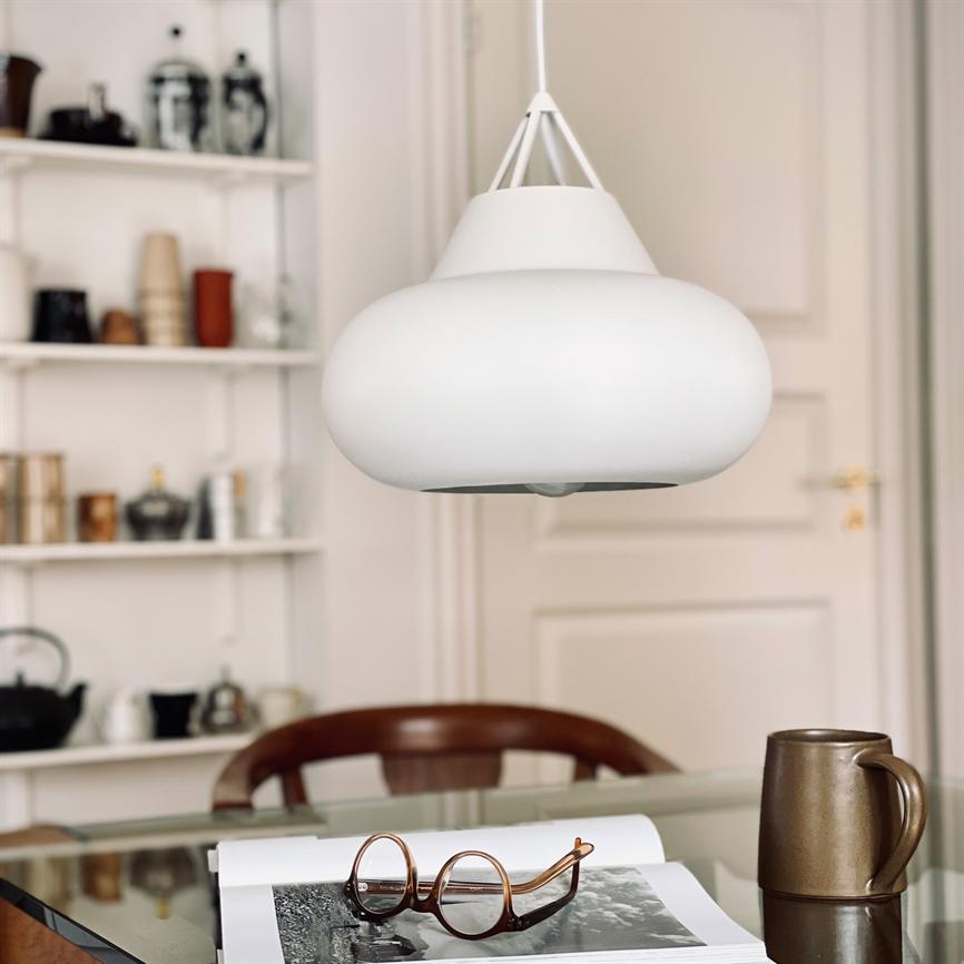 Hvid loftslampe hænger over et glasbord; på bordet ligger en åben bog med briller og et brunt krus; hylder med porcelæn og en stol i baggrunden.