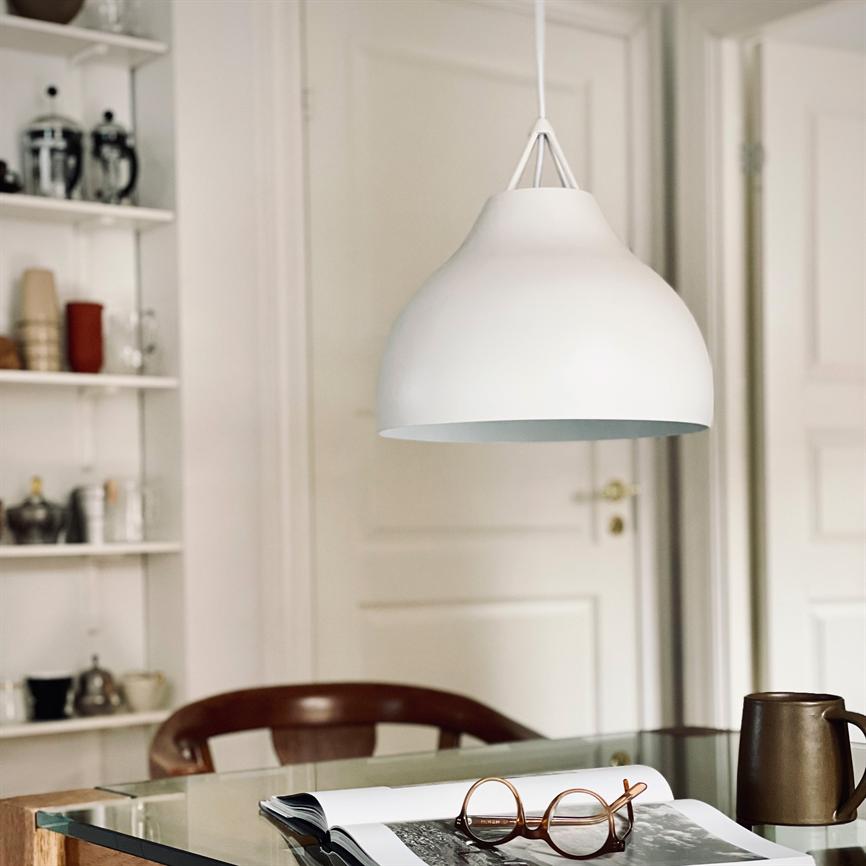 En hvid pendellampe hænger over et glasbord; et åbent magasin med brune briller og en brun krus ligger på bordet, med en stol og hylde i et lyst rum.