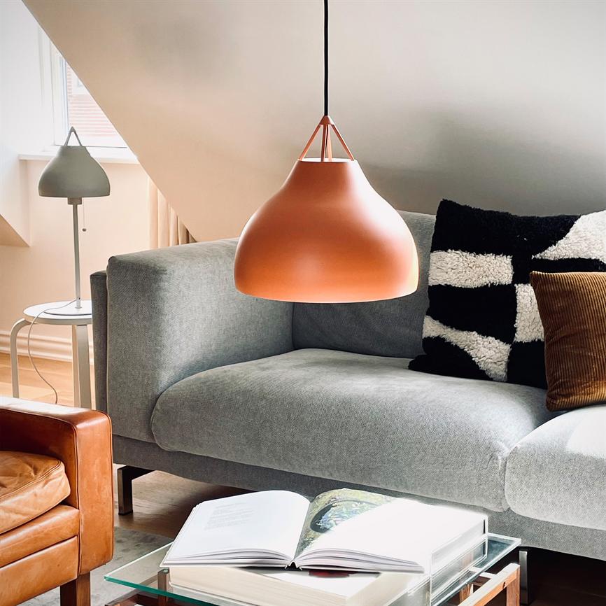 En orange pendellampe hænger over en åben bog på et glas sofabord; en grå sofa med sort-hvidt mønstret pude og brun fløjlsagtig pude står i et lyst, moderne stuehjørne.