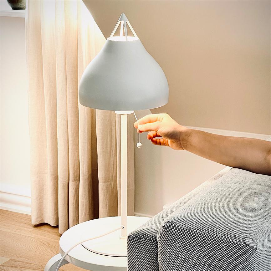 Hvid bordlampe, en hånd trækker tændesnoren; lampen står på et rundt sidebord ved en lysegrå sofa foran cremefarvede gardiner i et lyst stuerum.