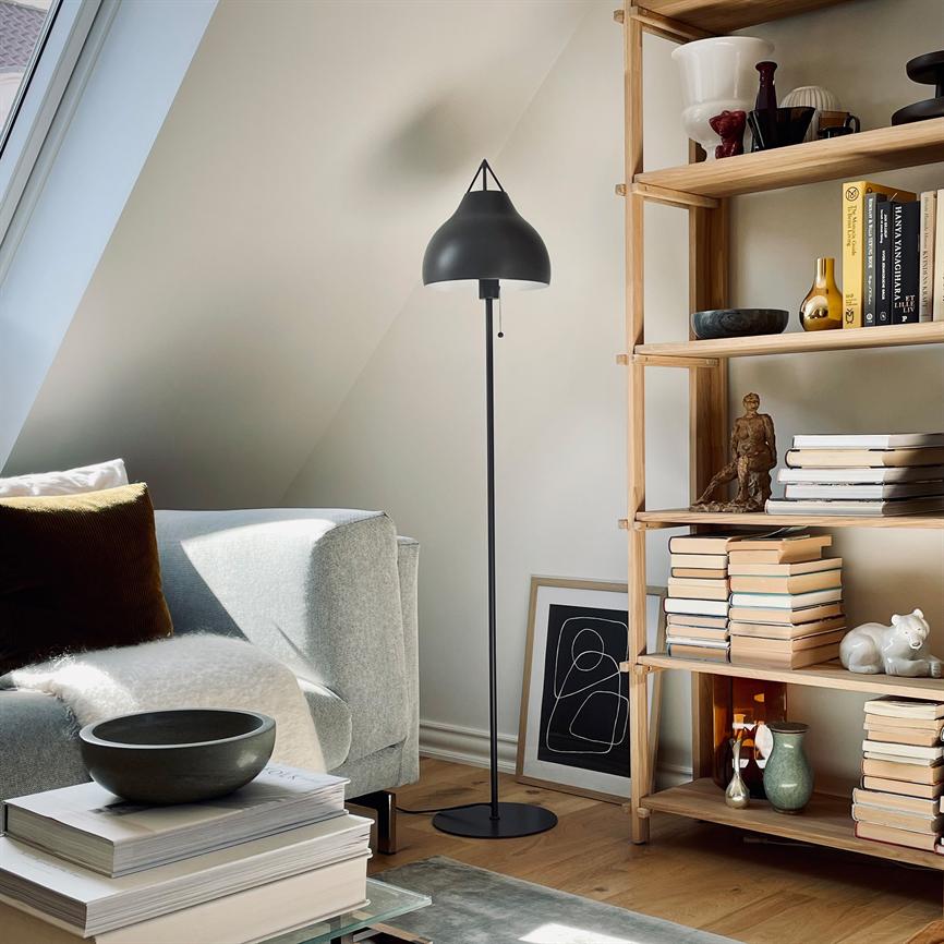Gulvlampe står oprejst mellem en lys sofa og en åben bogreol i en solbelyst taglejlighed, mens bøger, skåle og en indrammet plakat pryder gulvet og hylderne.
