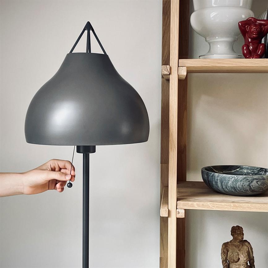 Grafitgrå gulvlampe trukket i snoren af en hånd, placeret ved en lys egetræshylde med en marmor skål, keramisk vase og en lille skulptur mod en hvid væg.