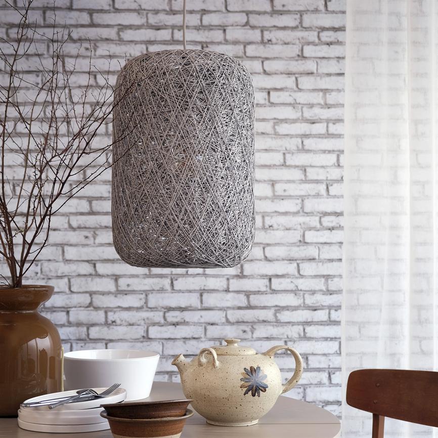 En vævet cylindrisk loftslampe hænger over et bord, hvor en keramisk tekande, skåle, bestik og en vase med nøgne grene står foran en hvid murstensvæg og let gardin.