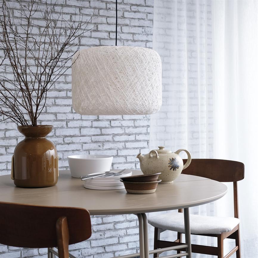 Stor hvid tekstureret loftlampe hænger over et rundt spisebord; en keramikkande, skåle og en brun vase med grene står på bordet i et lyst hjørne med hvidt murstensvæg og tylgardin.