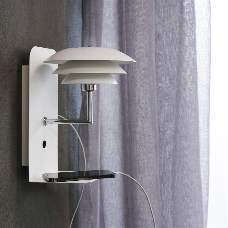Væglampe med lagdelt skærm holder en lille hylde, hvor en smartphone oplades via synligt kabel; monteret på væg foran et blødt, lysegråt gardin i et moderne rum.