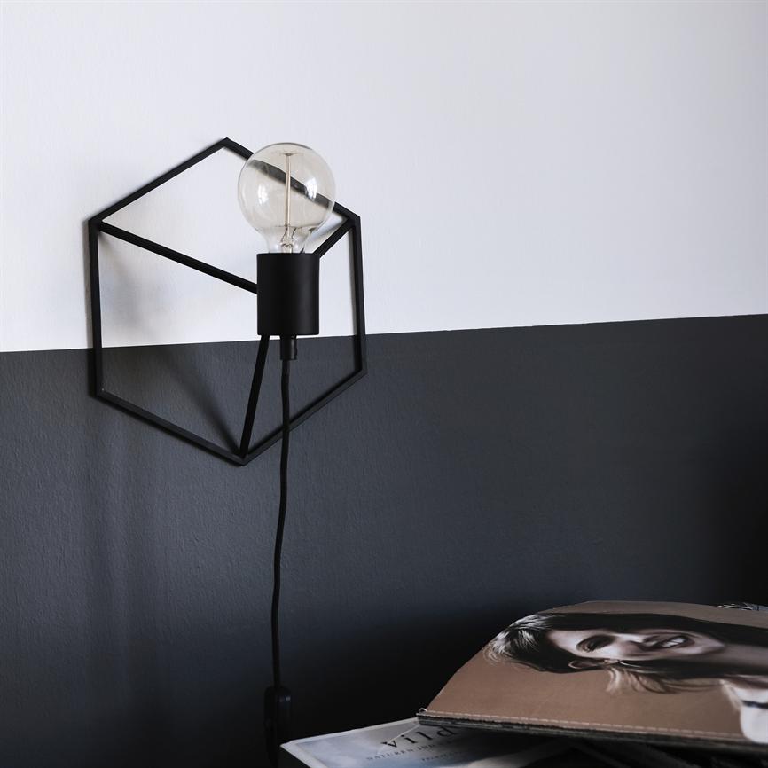Geometrisk sort metalvæglampe bærer en klar glødepære; den er monteret på en todelt hvid‑og‑mørkegrå væg over en overflade, hvor et magasin med et portræt af en kvinde ligger.