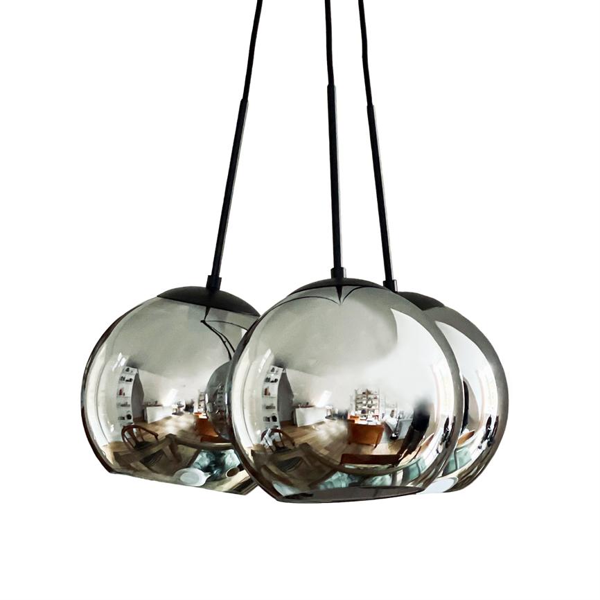 Tre spejlblanke, kugleformede pendellamper hænger fra sorte ledninger og spejler et lyst, moderne rum med møbler, hylder og trægulv.