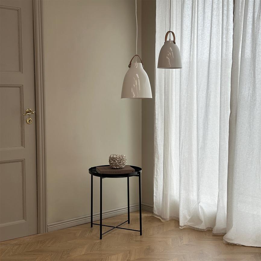 Et lille rundt sort sidebord står i et hjørne og bærer en bog samt en hvid korallignende dekoration under to hvide pendellamper foran let gennemskinnelige gardiner og en beige væg.