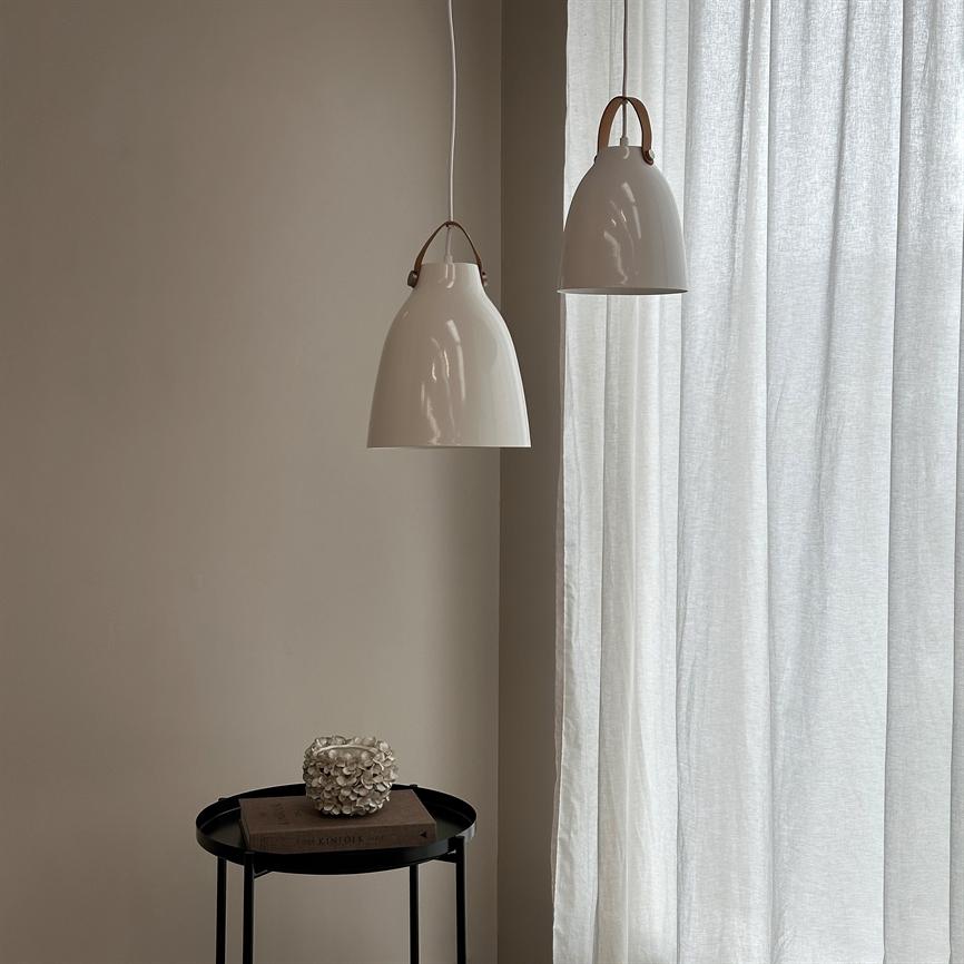 To hvide pendellamper hænger over et lille sort sidebord, som bærer en bog og en dekorativ hvid genstand, foran lyse, gennemsigtige gardiner i et neutralt beige rum.