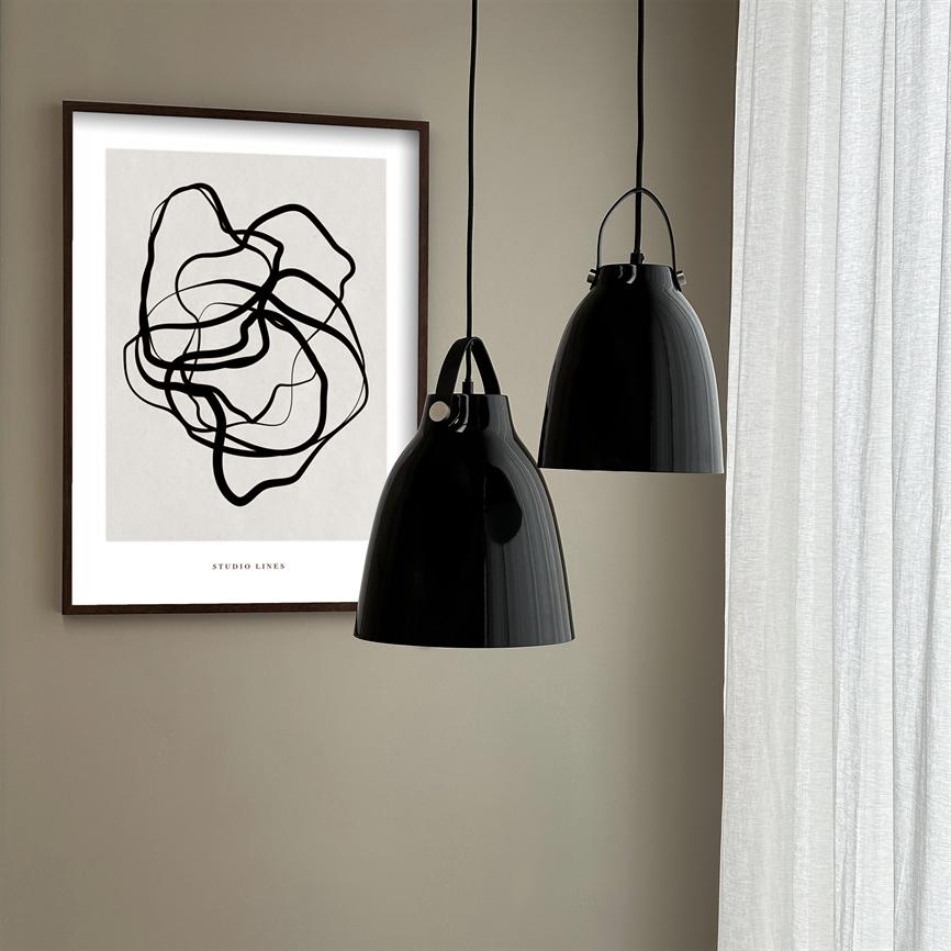 Et sort-hvidt abstrakt plakatmotiv viser sammenfiltrede sorte linjer; teksten 'STUDIO LINES' står nederst. Motivets ramme hænger ved siden af to blanke sorte pendellamper foran lyse gardiner i et neutralt rum.