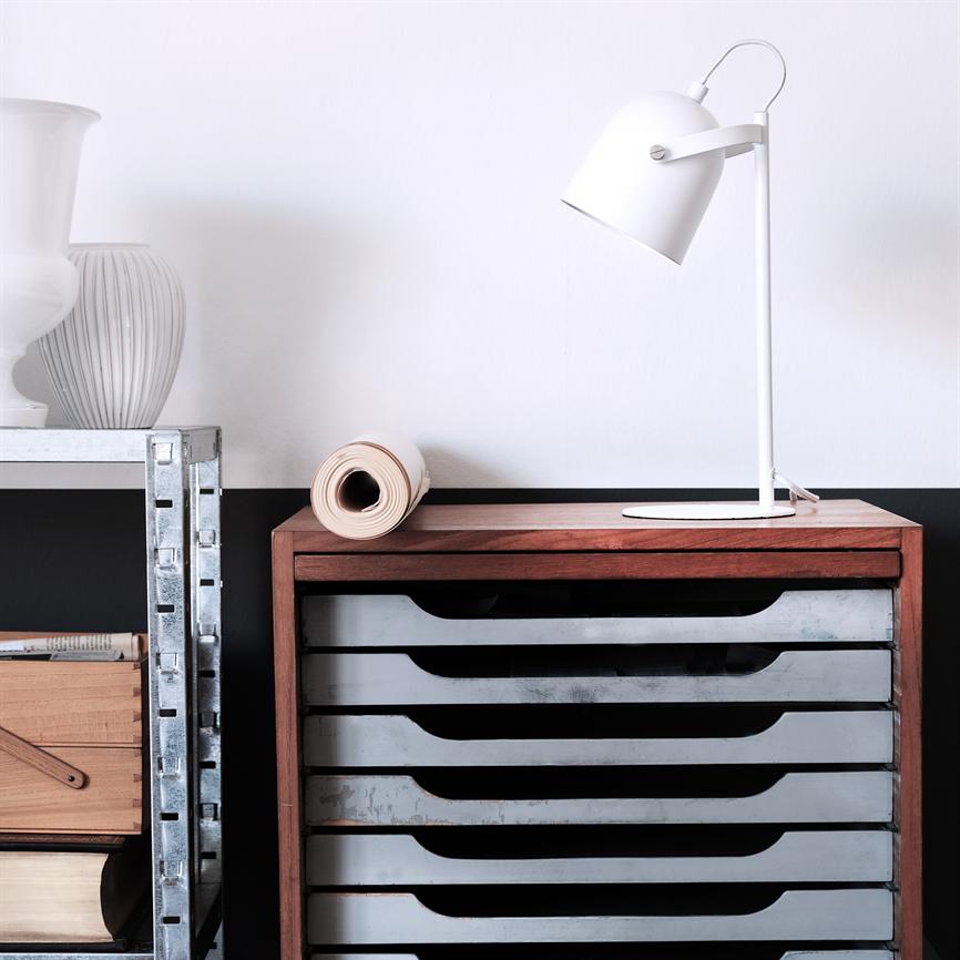 Hvid bordlampe står vinklet på et træskab med metalbakker; et sammenrullet papir ligger ved siden af. Til venstre en metalhylde med vaser og trækasser i lyst, minimalistisk rum.